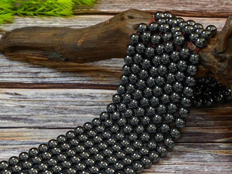 May include: A strand of black hematite beads, each bead is round and smooth. The beads are strung on a thin cord and are displayed on a piece of driftwood.