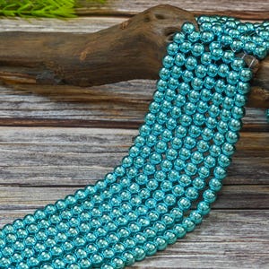 May include: A strand of turquoise blue metallic beads. The beads are round and have a shiny finish. The beads are strung on a clear thread and are lying on a wooden surface.