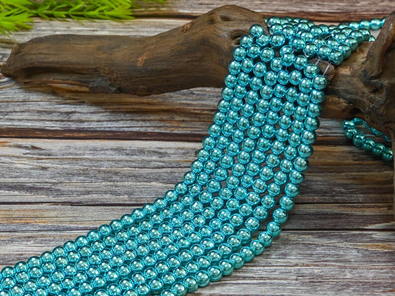 May include: A strand of turquoise blue metallic beads. The beads are round and have a shiny finish. The beads are strung on a clear thread and are lying on a wooden surface.
