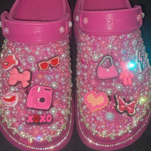Op de afbeelding: Een paar felroze Crocs-sandalen, rijkelijk versierd met parels, strass-steentjes en diverse bedels. Bedels zijn onder meer een camera, een strik, een zonnebril en een handtas. De sandalen zijn versierd met de tekst "XOXO".