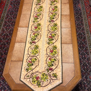May include: A long, rectangular quilted table runner featuring a cream-colored background with a repeating pattern of brown winding vines, green grape leaves, and clusters of red and green grapes, finished with a dark green fabric border.