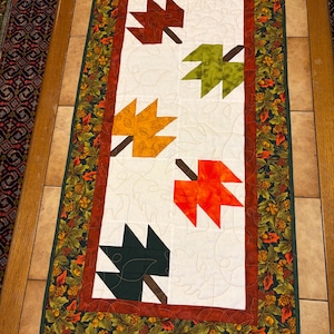 May include: A rectangular quilted table runner featuring a white center panel with five appliqued autumn leaves in shades of dark red, olive green, gold, orange, and dark green. The runner has a rust-colored inner border and a dark green outer border.