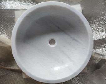 May include: White marble sink with a round shape and a small drain hole in the center.