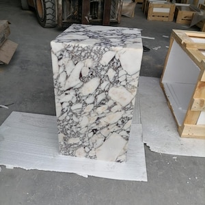 May include: A white marble cube with a purple and gray veining pattern. The cube is sitting on a white surface.