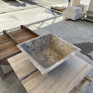 May include: A square, light brown stone planter with a rough texture. The planter is sitting on a wooden platform with a light brown stone surface.