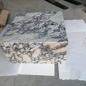 May include: A rectangular marble block with a striking pattern of white, gray, and purple veining. The stone is cut into a cube shape, showcasing the natural variations in color and texture. This decorative piece could be used as a coffee table or accent piece.