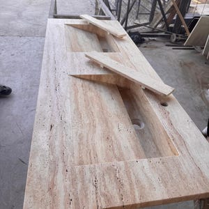 May include: A double sink vanity countertop made of travertine stone. The countertop has a light beige color with brown streaks and a natural, textured finish.