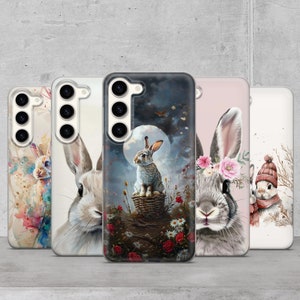 May include: Five different phone cases featuring watercolour illustrations of rabbits. The cases are all clear and show the phone's camera lens. The rabbits are depicted in various poses and settings, including a rabbit sitting in a basket under a full moon.