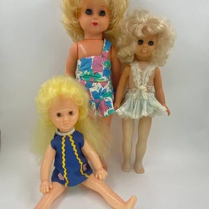 Könnte beinhalten: Drei Vintage-Plastikpuppen mit blonden Haaren. Die Puppen tragen verschiedene Outfits. Eine Puppe trägt ein blaues und gelbes Kleid, eine andere trägt ein Blumenkleid und die dritte trägt ein weißes Spitzenkleid.