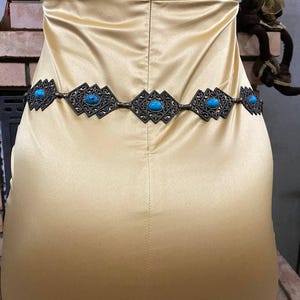 May include: A gold-toned belt with turquoise stones and a geometric design. The belt is worn around the waist of a person wearing a gold satin dress.
