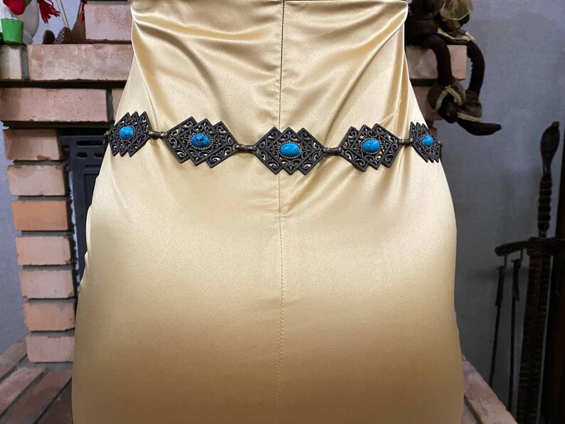 May include: A gold-toned belt with turquoise stones and a geometric design. The belt is worn around the waist of a person wearing a gold satin dress.