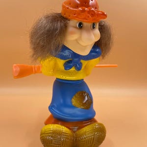 Può includere: Statuetta di strega in plastica vintage. La strega ha un viso sorridente, capelli castani e indossa un cappello arancione, una giacca gialla, un vestito blu e scarpe marroni. Tiene una scopa arancione.