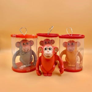 May include: Three toy monkeys in clear plastic containers with red lids. The monkeys are in different colors: gray, orange, and red. The red monkey is outside of the container.