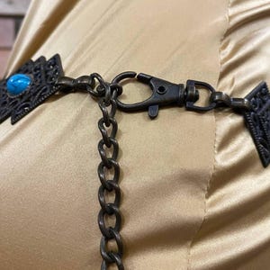 May include: A close-up of a brown chain belt with turquoise stones and a metal clasp. The belt is on a light brown fabric background.
