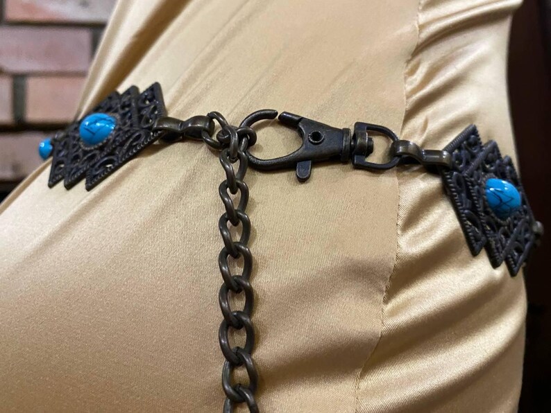 May include: A close-up of a brown chain belt with turquoise stones and a metal clasp. The belt is on a light brown fabric background.