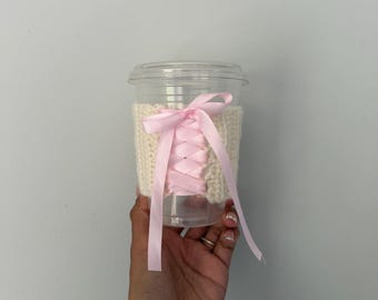 May include: A clear plastic cup featuring a cream-colored crocheted sleeve. The sleeve is decorated with a light pink satin ribbon laced in a crisscross corset pattern, tied into a bow at the top center, providing a textured and layered appearance.