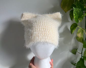 May include: A cream-colored, fuzzy knit hat with pointed ear details. The hat is displayed on a white mannequin head. The hat is held by a hand with a silver bracelet. The background is a light gray wall with green plants.