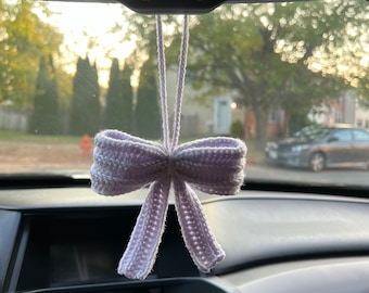 May include: A crocheted bow car charm in shades of lavender and gray hangs from a car's rearview mirror. The bow is suspended by two matching cords. The background shows a residential street with trees and a car.