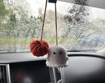 May include: A hanging car accessory featuring a small, crocheted orange pumpkin and a white ghost with black bead eyes. Both items are suspended by thin, light brown twine strings from a rearview mirror, creating a decorative pair.