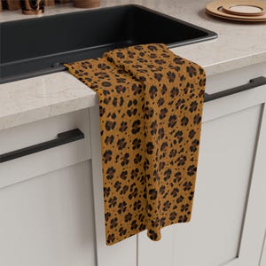 May include: A rectangular kitchen towel featuring a repeating leopard print pattern in shades of golden brown, dark brown, and black. The fabric has a textured weave and is draped over the edge of a white kitchen counter next to a black sink.