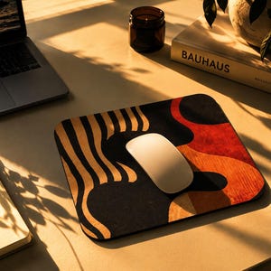 Könnte beinhalten: Ein rechteckiges Mauspad mit einem abstrakten Design aus schwarzen, roten und orangefarbenen wellenförmigen Formen und Streifen. Eine glatte, weiße, kabellose Computermaus befindet sich in der Mitte des Pads auf der gemusterten Oberfläche.