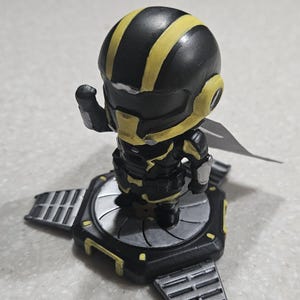 May include: A small figurine of a character in a black and yellow armored suit with a helmet. The figure is standing on a black and silver platform with three rectangular extensions. The figure has a wing-like element on its back.