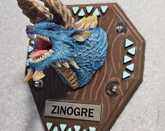 May include: A detailed, blue and yellow dragon head mount with open mouth and visible teeth. The mount is attached to a brown, hexagonal plaque with decorative triangles and the name "ZINOGRE" on a silver nameplate.