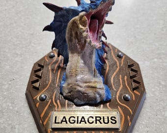 May include: A detailed, hand-painted sculpture of a Lagiacrus monster head, featuring blue, red, and tan colors. The sculpture is mounted on a brown, hexagonal plaque with the name "LAGIACRUS" displayed prominently.