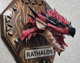 May include: A detailed, hand-painted dragon head sculpture with red, black, and beige tones, mounted on a brown hexagonal plaque. The plaque features the name "RATHALOS" and decorative elements.