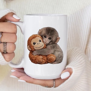 May include: White ceramic mug featuring a charming illustration of two hugging monkeys. One monkey is brown and the other is gray. The mug has a large handle and is held by a person wearing a white sweater.