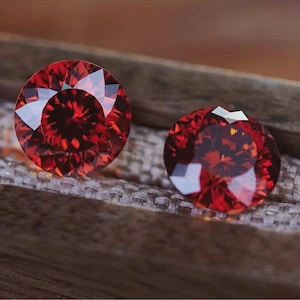 Peut inclure: Deux pierres précieuses rondes, facettées, d'un rouge profond. Les gemmes sont présentées sur une surface texturée de tons neutres, avec un élément en bois en arrière-plan. Les gemmes ont une taille brillante, reflétant la lumière et mettant en valeur leur couleur riche.