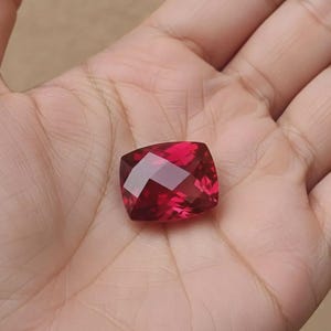 May include: A faceted, square-cut, deep red gemstone. The gem is held in a hand, showcasing its brilliant, reflective surfaces. The stone's color is a rich, saturated red, with multiple facets catching the light.