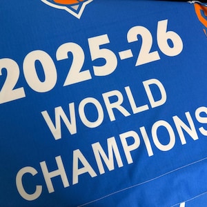 May include: A bright blue fabric banner featuring the white text 2025-26 WORLD CHAMPIONS. The design includes partial orange and white sports team logos at the top corners, printed on a smooth, solid-coloured textile material.