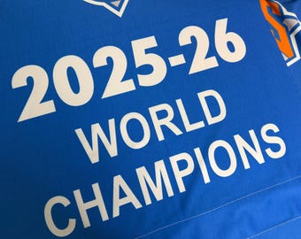 May include: A bright blue fabric banner featuring the white text 2025-26 WORLD CHAMPIONS. The design includes partial orange and white sports team logos at the top corners, printed on a smooth, solid-colored textile material.