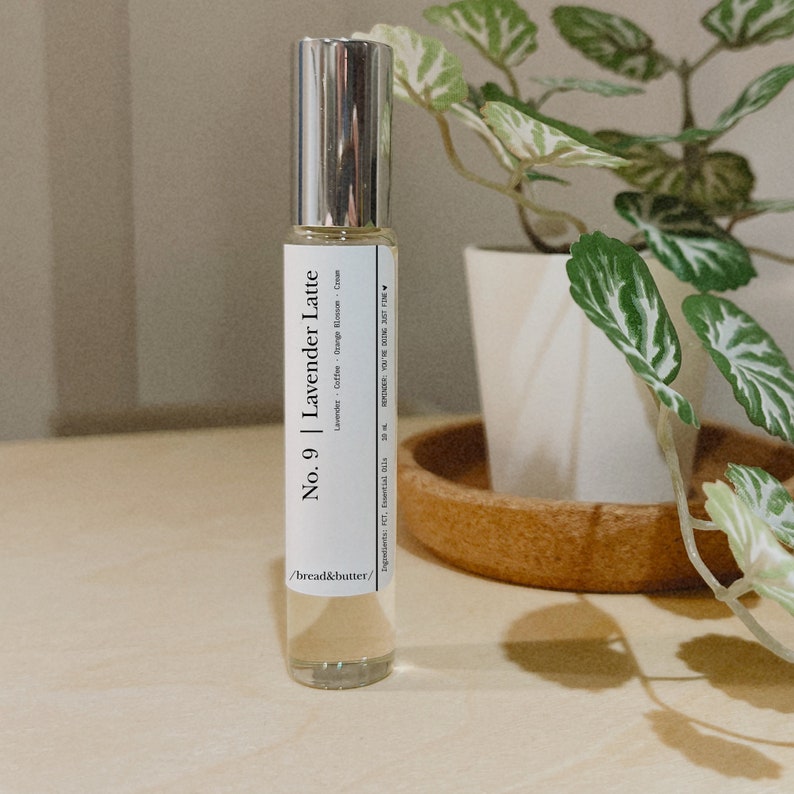 May include: A clear glass roll-on bottle with a silver cap. The label reads "No. 9 | Lavender Latte" with ingredients listed below. The bottle is on a wooden surface, with a plant in the background.
