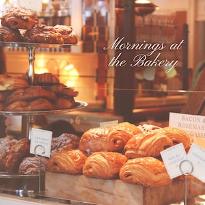 Può includere: Un'esposizione di panetteria con vari pasticcini, tra cui croissant e pain au chocolat. Il testo "Mornings at the Bakery" è visibile. I prodotti da forno sono presentati su vassoi d'argento e superfici in legno.