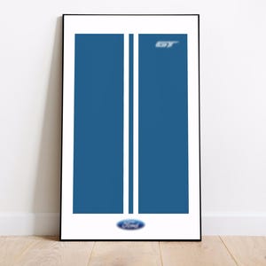 以下が含まれることがあります： 青色の背景に2本の白い縦縞が入った額入りプリント。右上隅には銀色の文字で「GT」と表示され、下部にはフォードのロゴがあります。アートワークは黒いフレームで表示されています。