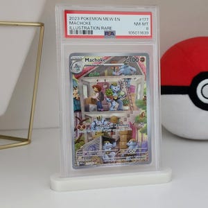 Puede incluir: Una carta Pokémon Machoke del set 2023 Mew EN, clasificada y protegida en una funda transparente. La carta está etiquetada como "Machoke Illustration Rare". La ilustración muestra a Machoke en un edificio de varios niveles. Una Poké Ball roja y blanca en el fondo.