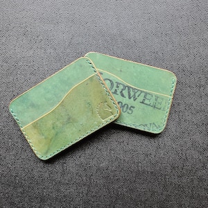 May include: Two green leather cardholders with stitched edges. One cardholder has a lighter green pocket and a stamped logo. The other cardholder has the word "ORWELL" and the number "05" stamped on it.