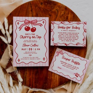 May include: Pink baby shower invitations and inserts with a cherry theme. The main invitation reads "She's the Cherry on Top" and includes event details. Additional cards request books and announce a diaper raffle.