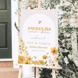 Puede incluir: Un letrero de bienvenida en un caballete con el texto "sweet as can bee" y "Kat & Luke's babee shower". El letrero presenta un patrón de panal, ilustraciones florales amarillas y gráficos de abejas. También se muestra la fecha 15 de junio.