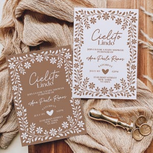 May include: Two baby shower invitations with floral borders and the text "Cielito Lindo." The invitations are in brown and white, with the details of the event. A gold wax seal and coins are on the side.