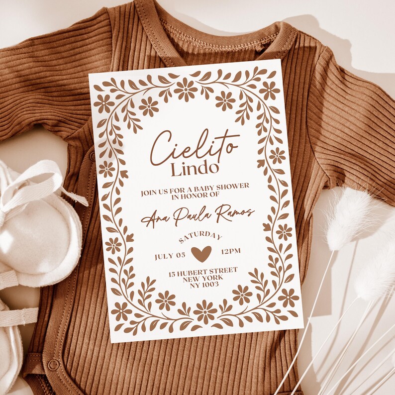 May include: A baby shower invitation with the words "Cielito Lindo" in a decorative font, surrounded by a floral border. The invitation is on a white background and includes the name "Ana Paula Ramos", date, time, and address. The invitation is placed on a brown onesie.