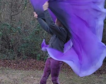 May include: A person is holding a large, flowing purple flag outdoors. The flag has a gradient effect, transitioning from a darker shade at the bottom to a lighter purple at the top. The person is wearing a green jacket, purple leggings, and black boots.