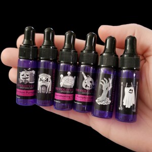 May include: Seven small purple glass bottles with black dropper tops and labels featuring Halloween-themed illustrations. The labels include text such as "The Violet Vul Apothecary", "Pumpkin Candle", and "Summoning Circle".