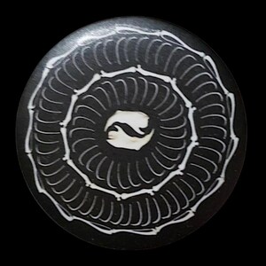 May include: A round, black object with a white spiral design. The spiral is composed of curved lines, and the center features a white, abstract shape. The object has a smooth surface and is set against a black background.