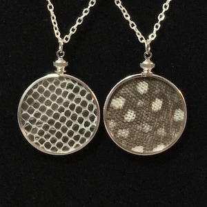 May include: Two silver necklaces with round pendants. The pendants are clear and have a silver frame. One pendant has a white and gray patterned design, and the other has a white honeycomb pattern.
