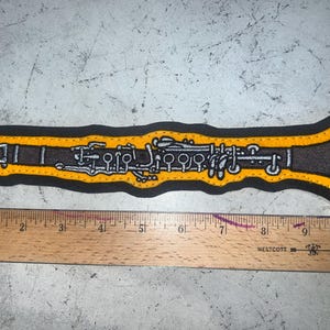 May include: A yellow and black embroidered patch of a clarinet. The clarinet is outlined in black and has a yellow body with black and white details. The patch is approximately 6 inches long.