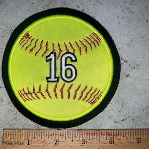 May include: A round, yellow softball patch with red stitching and the number 16 in white with a black outline. The patch has a black border. A wooden ruler is at the bottom of the image.