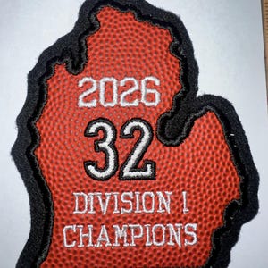 Puede incluir: Parche bordado con la forma del estado de Michigan, con textura de baloncesto rojo. El parche tiene el texto blanco "2026 32 DIVISION 1 CHAMPIONS", con un borde negro.